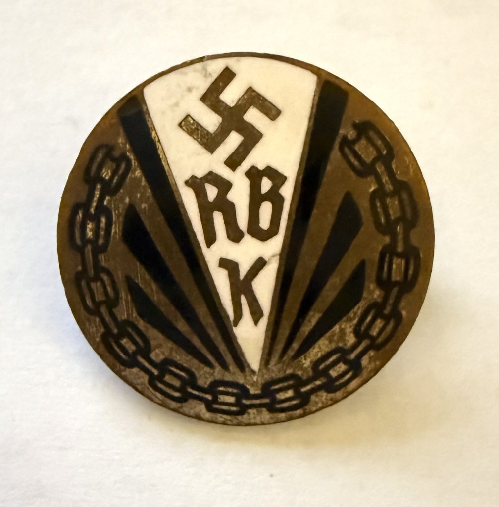 Rundes Abzeichen aus Messing. In der Mitte ein Dreieck mit weißem Hintergrund, darauf ein Hakenkreuz und RBK in Messingfarbe. An den Seiten je zwei schwarze Striche und eine Kette in schwarzer Farbe.