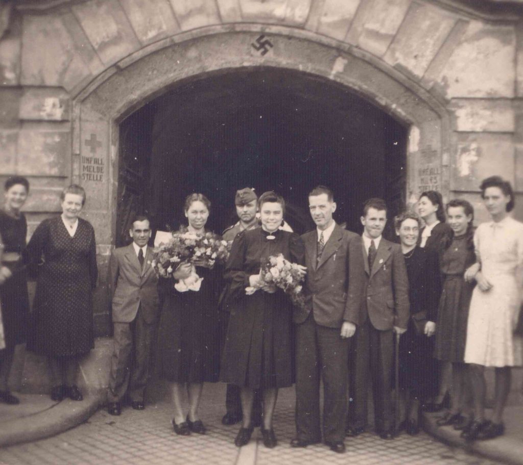 Eine Hochzeitsgesellschaft mit Menschen ohne sichtbare Behinderungen vor dem Rathaus in St. Pölten. Über dem Eingangstor ein Hakenkreuz.