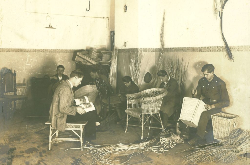 In einer Werkstatt verrichten fünf Männer Korbflechtarbeiten. Ein Mann sitzt in der Mitte und liest ein Buch. Das Bild wirkt arrangiert.