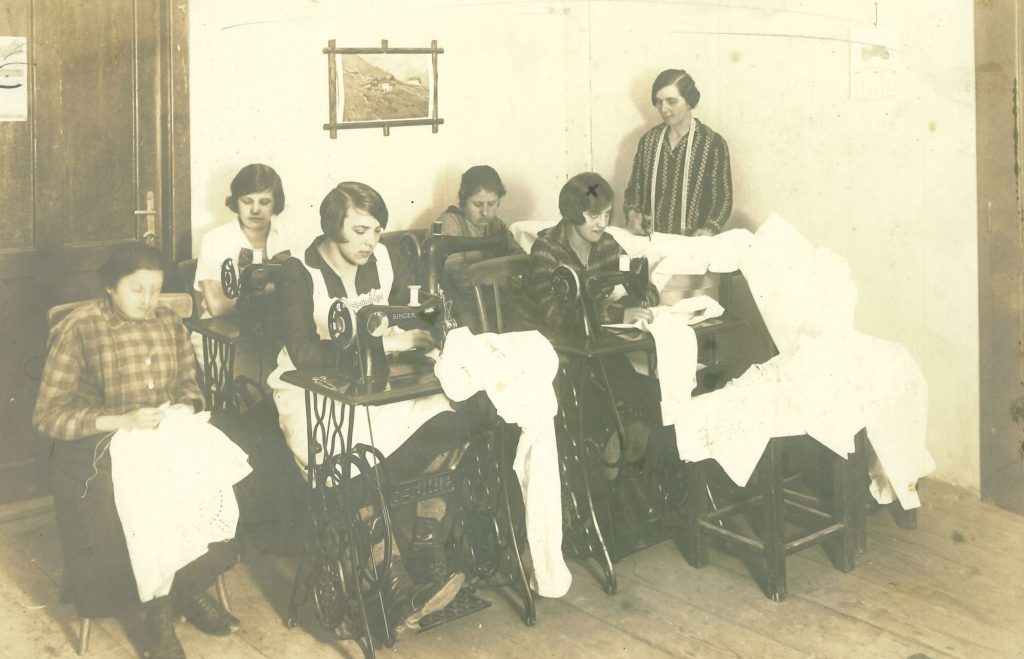 Auf dem Foto sind sechs Frauen aus der Nähwerkstatt mit ihren Textilarbeiten zu sehen. Vier davon sitzen an Nähmaschinen. Das Foto wirkt arrangiert.