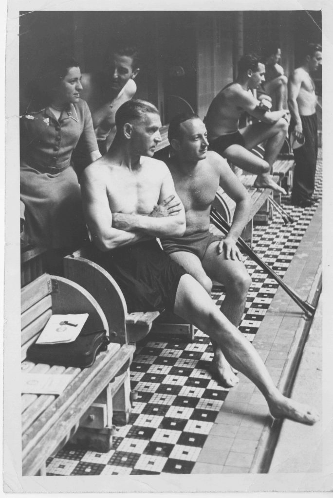 Im Schwimmbad: Vorne in Badehose der sportlich junge Wilfried Kobras. Im Hintergrund sind mehr Männer in Badehosen zu sehen. Hinter Kobras in Kleidung die junge Grete Ruckstätter.