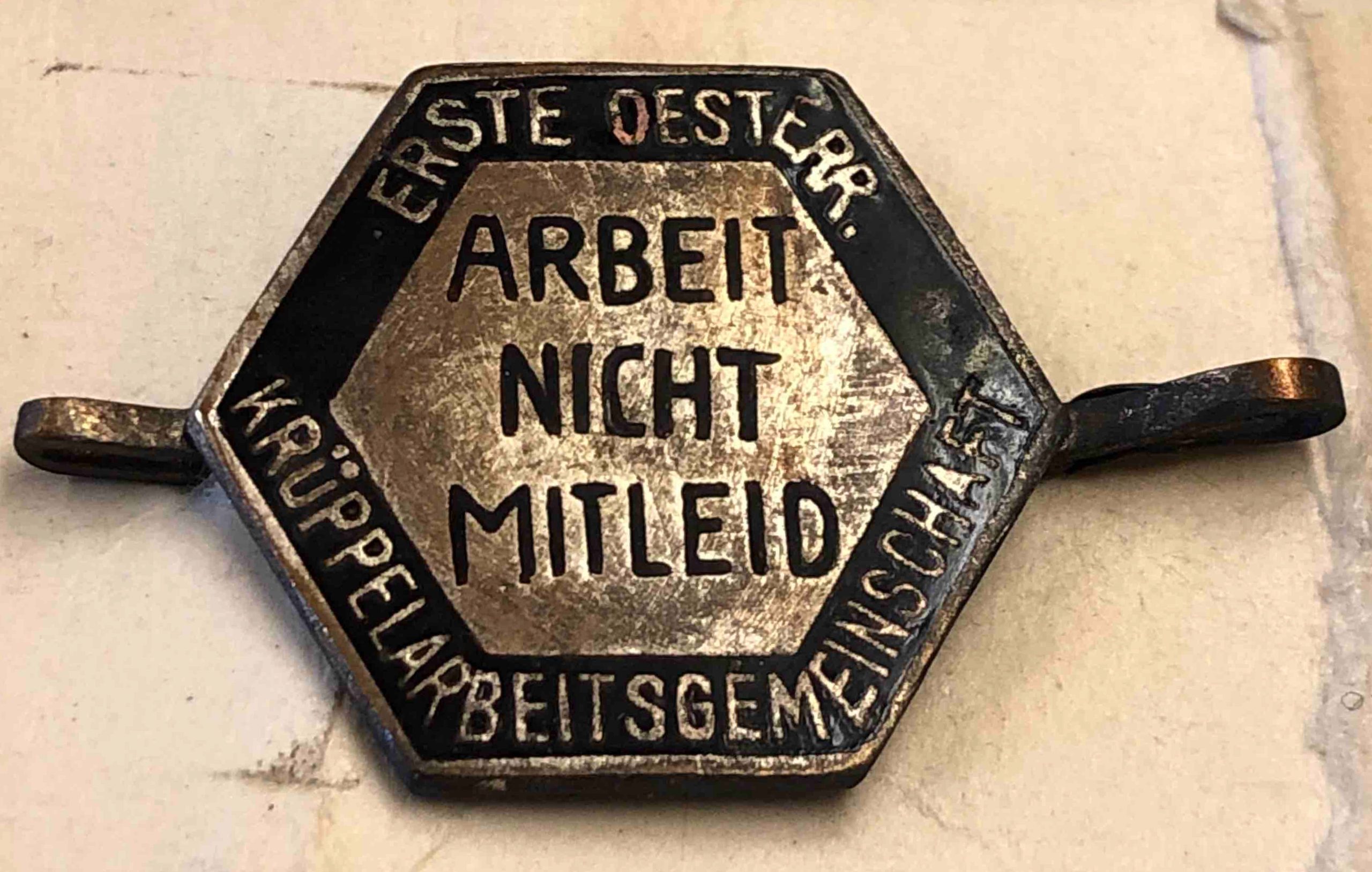 Auf dem regelmäßigen sechseckigen Abzeichen (Breite: 1,9 cm, mit Nadel zur Befestigung: 3,3 cm, Höhe: 1,7 cm) steht in der Mitte auf einem messingfarbenen Hintergrund mit schwarzer Schrift „ARBEIT NICHT MITLEID“. Im schwarzen Rahmen steht in heller Schrift „ERSTE OESTERR. KRÜPPELARBEITSGEMEINSCHAFT“.